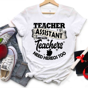 K&ouml;nnte beinhalten: Wei&szlig;es T-Shirt mit schwarzem Text, der "Teacher Assistant Because Teachers Need Heros Too" lautet. Der Text ist von einem schwarzen Band mit Sternen umgeben. Ein roter Apfel mit einem Bissen ist in der Mitte des Textes.
