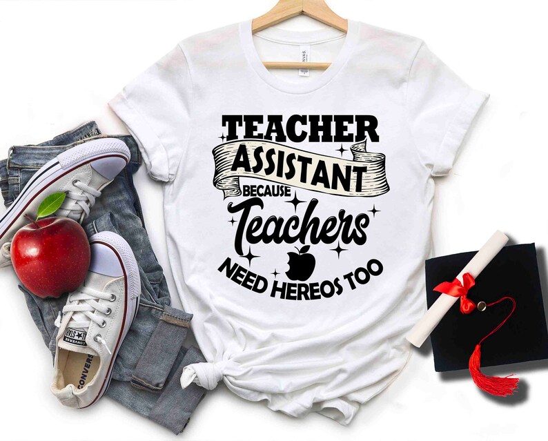 K&ouml;nnte beinhalten: Wei&szlig;es T-Shirt mit schwarzem Text, der "Teacher Assistant Because Teachers Need Heros Too" lautet. Der Text ist von einem schwarzen Band mit Sternen umgeben. Ein roter Apfel mit einem Bissen ist in der Mitte des Textes.