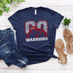 Puede incluir: Una camiseta azul marino con un gr&aacute;fico de baloncesto blanco y rojo. El bal&oacute;n de baloncesto est&aacute; delineado en rojo y tiene un dise&ntilde;o de estampado de leopardo blanco. El texto "WARRIORS" est&aacute; impreso en blanco debajo del bal&oacute;n de baloncesto.