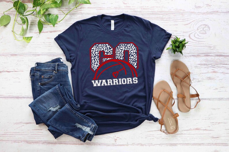 Puede incluir: Una camiseta azul marino con un gr&aacute;fico de baloncesto blanco y rojo. El bal&oacute;n de baloncesto est&aacute; delineado en rojo y tiene un dise&ntilde;o de estampado de leopardo blanco. El texto "WARRIORS" est&aacute; impreso en blanco debajo del bal&oacute;n de baloncesto.