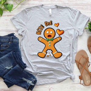 Op de afbeelding: Een grijs T-shirt met een afbeelding van een peperkoekman en de tekst "Bite Me" in wit. De peperkoekman heeft een verraste uitdrukking en houdt een rood hart vast.