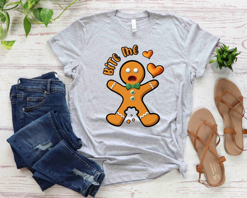 Op de afbeelding: Een grijs T-shirt met een afbeelding van een peperkoekman en de tekst "Bite Me" in wit. De peperkoekman heeft een verraste uitdrukking en houdt een rood hart vast.