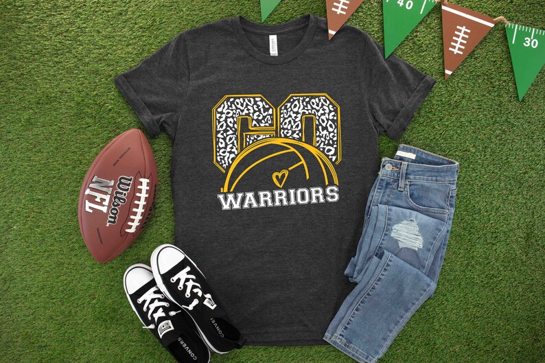 Puede incluir: Una camiseta gris oscuro con un gr&aacute;fico de baloncesto dorado y blanco. El bal&oacute;n de baloncesto tiene un coraz&oacute;n blanco en el centro. El texto "GO WARRIORS" est&aacute; impreso debajo del bal&oacute;n de baloncesto.
