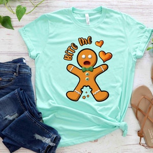 Op de afbeelding: Een lichtblauw t-shirt met een afbeelding van een peperkoekman en de tekst "Bite Me" met een hart. De peperkoekman draagt een groene strik en heeft een verraste uitdrukking.