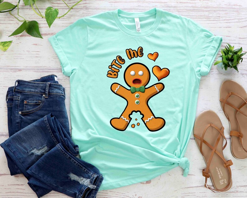 Op de afbeelding: Een lichtblauw t-shirt met een afbeelding van een peperkoekman en de tekst "Bite Me" met een hart. De peperkoekman draagt een groene strik en heeft een verraste uitdrukking.