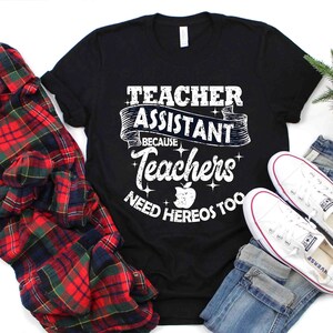 K&ouml;nnte beinhalten: Schwarzes T-Shirt mit wei&szlig;em Text, der "Teacher Assistant Because Teachers Need Heros Too" lautet. Der Text ist in einer verwitterten Schriftart mit einem roten Apfel-Motiv. Das Shirt liegt auf einem karierten Hemd, einer blauen Jeans und wei&szlig;en Turnschuhen.