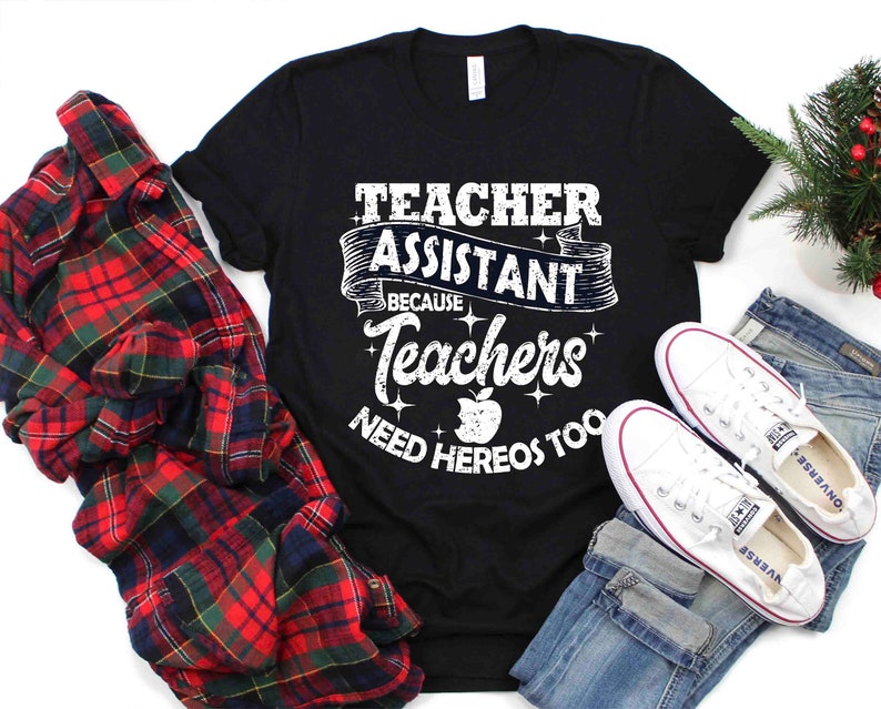 K&ouml;nnte beinhalten: Schwarzes T-Shirt mit wei&szlig;em Text, der "Teacher Assistant Because Teachers Need Heros Too" lautet. Der Text ist in einer verwitterten Schriftart mit einem roten Apfel-Motiv. Das Shirt liegt auf einem karierten Hemd, einer blauen Jeans und wei&szlig;en Turnschuhen.
