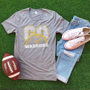 Puede incluir: Una camiseta gris con un dise&ntilde;o blanco y dorado que presenta el n&uacute;mero "60" con un bal&oacute;n de baloncesto y las palabras "WARRIORS". La camiseta est&aacute; colocada sobre una superficie verde con un bal&oacute;n de f&uacute;tbol, unos vaqueros y unas zapatillas blancas.