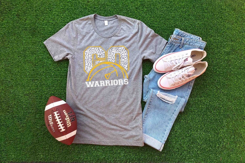 Puede incluir: Una camiseta gris con un dise&ntilde;o blanco y dorado que presenta el n&uacute;mero "60" con un bal&oacute;n de baloncesto y las palabras "WARRIORS". La camiseta est&aacute; colocada sobre una superficie verde con un bal&oacute;n de f&uacute;tbol, unos vaqueros y unas zapatillas blancas.