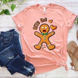 Op de afbeelding: Een roze T-shirt met een afbeelding van een peperkoekman en de tekst "Bite Me" in wit. De peperkoekman draagt een groene strik en heeft een verraste uitdrukking. Er zijn twee rode harten boven de peperkoekman.