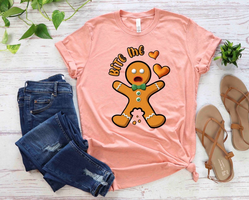 Op de afbeelding: Een roze T-shirt met een afbeelding van een peperkoekman en de tekst "Bite Me" in wit. De peperkoekman draagt een groene strik en heeft een verraste uitdrukking. Er zijn twee rode harten boven de peperkoekman.