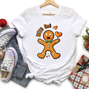 Op de afbeelding: Wit T-shirt met een afbeelding van een peperkoekman en de tekst "Bite Me" in rood en zwart. De peperkoekman draagt een groene strik en heeft een verraste uitdrukking. Boven de peperkoekman staan twee rode harten.