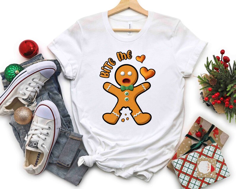 Op de afbeelding: Wit T-shirt met een afbeelding van een peperkoekman en de tekst "Bite Me" in rood en zwart. De peperkoekman draagt een groene strik en heeft een verraste uitdrukking. Boven de peperkoekman staan twee rode harten.