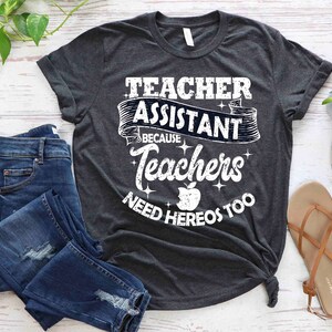 K&ouml;nnte beinhalten: Ein dunkelgraues T-Shirt mit einer wei&szlig;en Grafik, auf der "Teacher Assistant Because Teachers Need Heros Too" steht. Die Grafik zeigt einen roten Apfel und ein Banner mit den Worten "Teacher Assistant" in einer Vintage-Schriftart.