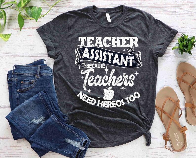 K&ouml;nnte beinhalten: Ein dunkelgraues T-Shirt mit einer wei&szlig;en Grafik, auf der "Teacher Assistant Because Teachers Need Heros Too" steht. Die Grafik zeigt einen roten Apfel und ein Banner mit den Worten "Teacher Assistant" in einer Vintage-Schriftart.