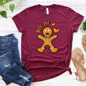 Op de afbeelding: Een maroon t-shirt met een afbeelding van een peperkoekman en de tekst "Bite Me" in wit. De peperkoekman draagt een groene strik en heeft een verraste uitdrukking. Er zijn twee rode harten boven de peperkoekman.