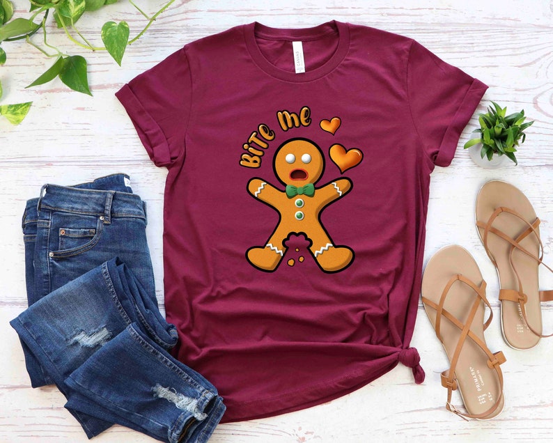 Op de afbeelding: Een maroon t-shirt met een afbeelding van een peperkoekman en de tekst "Bite Me" in wit. De peperkoekman draagt een groene strik en heeft een verraste uitdrukking. Er zijn twee rode harten boven de peperkoekman.