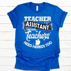 K&ouml;nnte beinhalten: Ein k&ouml;nigsblaues T-Shirt mit wei&szlig;em Text, der "Teacher Assistant Because Teachers Need Heros Too" lautet. Der Text ist in einer verwitterten Schriftart gehalten und enth&auml;lt eine kleine Apfelgrafik.