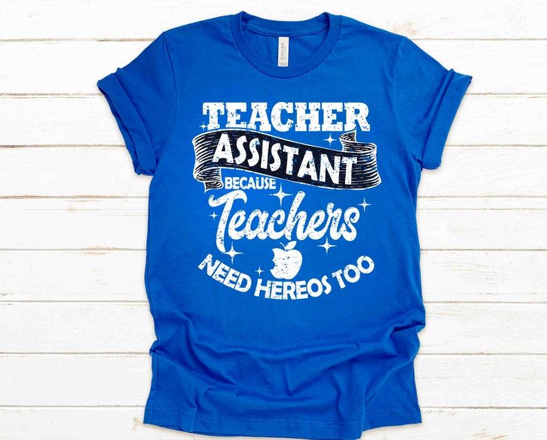 K&ouml;nnte beinhalten: Ein k&ouml;nigsblaues T-Shirt mit wei&szlig;em Text, der "Teacher Assistant Because Teachers Need Heros Too" lautet. Der Text ist in einer verwitterten Schriftart gehalten und enth&auml;lt eine kleine Apfelgrafik.