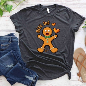 Op de afbeelding: Een donkergrijs T-shirt met een afbeelding van een peperkoekman en de tekst "Bite Me" in wit. De peperkoekman draagt een groene strik en heeft een verraste uitdrukking op zijn gezicht. Boven de tekst staan twee rode harten.