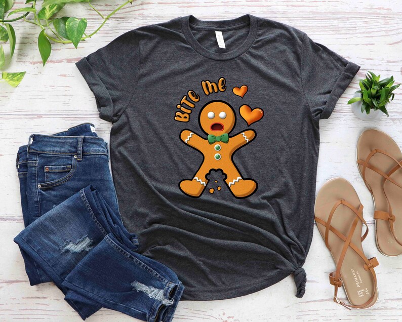 Op de afbeelding: Een donkergrijs T-shirt met een afbeelding van een peperkoekman en de tekst "Bite Me" in wit. De peperkoekman draagt een groene strik en heeft een verraste uitdrukking op zijn gezicht. Boven de tekst staan twee rode harten.
