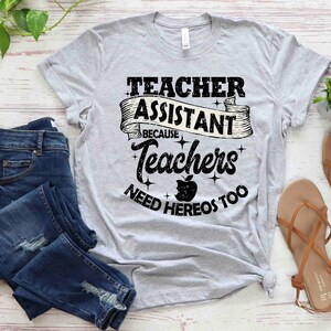 K&ouml;nnte beinhalten: Ein graues T-Shirt mit dem Text "Teacher Assistant Because Teachers Need Heros Too" in schwarzer Schrift mit einer schwarz-wei&szlig;en Apfelgrafik.