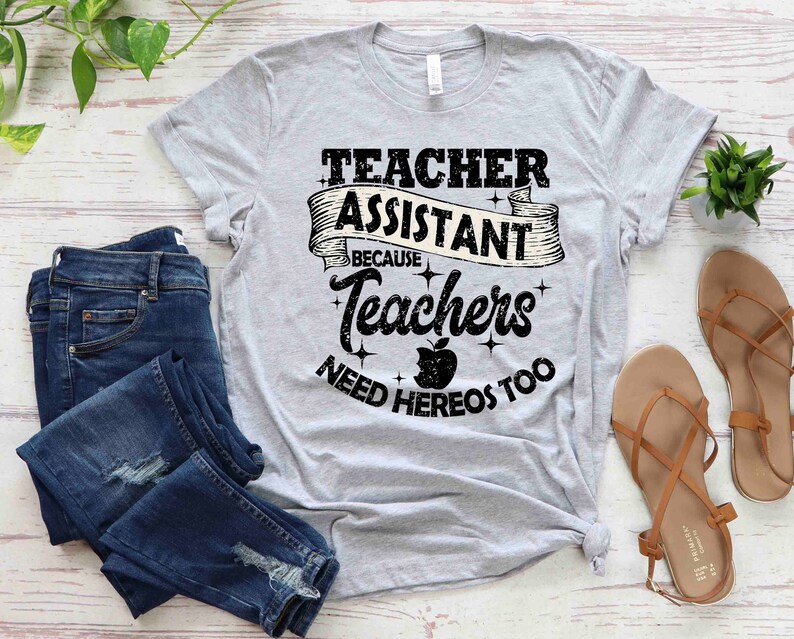 K&ouml;nnte beinhalten: Ein graues T-Shirt mit dem Text "Teacher Assistant Because Teachers Need Heros Too" in schwarzer Schrift mit einer schwarz-wei&szlig;en Apfelgrafik.