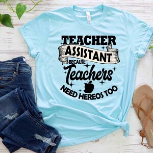 K&ouml;nnte beinhalten: Ein hellblaues T-Shirt mit dem Text "Teacher Assistant Because Teachers Need Heros Too" in Schwarz mit einer roten Apfelgrafik.