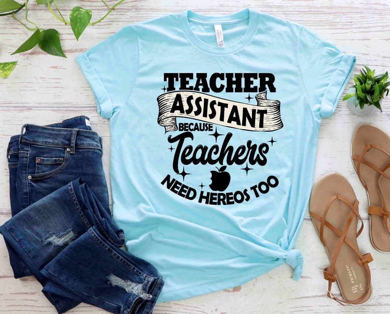 K&ouml;nnte beinhalten: Ein hellblaues T-Shirt mit dem Text "Teacher Assistant Because Teachers Need Heros Too" in Schwarz mit einer roten Apfelgrafik.