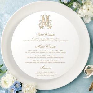 May include: A white plate with a menu for a wedding, featuring floral arrangements in shades of blue and white. The menu includes the courses: First Course, Main Course, and Dessert. Silverware is tied with a white ribbon.