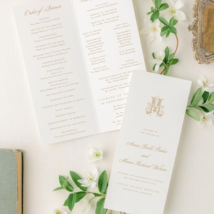 May include: Elegant wedding stationery featuring gold script on cream-colored paper. The image includes a tri-fold program, a smaller card with a monogram, and floral accents. The text includes names, dates, and the venue.