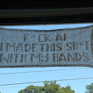Könnte beinhalten: Ein rechteckiger, dunkelgrauer Wandbehang aus Häkelarbeit mit dem Text F*CK AI I MADE THIS SH*T WITH MY HANDS. Das Stück ist in einem gitterartigen, offenen Maschenmuster gefertigt und hängt horizontal vor einem klaren, blauen Himmel.