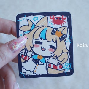 May include: A square sticker featuring a cartoon character with blonde hair, blue highlights, and cat ears. The character wears a white outfit with a blue bow and red-and-white striped accents. A small red crab is visible in the top right corner.