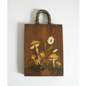 Puede incluir: Un colgador de pared de madera con un asa de cuerda presenta una escena pintada de setas y una flor blanca. Las setas son amarillas y marrones con tallos blancos. La flor es blanca con un centro amarillo.