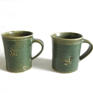 May include: Two green ceramic mugs with brown accents. Each mug has a different design of a bird with a branch in its beak.
