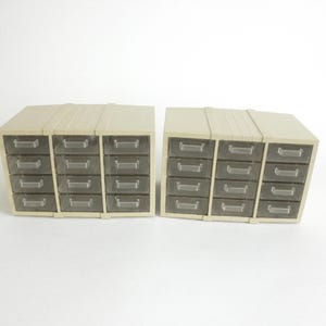 May include: Two beige plastic storage organizers, each featuring twelve small, clear drawers arranged in three vertical columns of four. The units have a ribbed top surface and integrated handles on each individual drawer for easy access.
