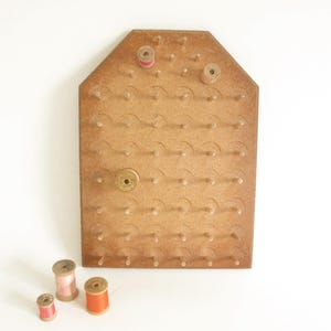 May include: A wooden thread spool holder with multiple pegs and a few spools of thread in pink and orange. The holder is a light brown color and has a unique shape.