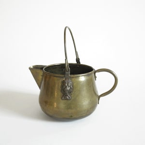 May include: Antique brass kettle with a spout and handle. The kettle has a rounded body, a curved handle, and a small spout. A decorative lion's head is attached to the handle's base. The kettle is made of brass and has a vintage appearance.