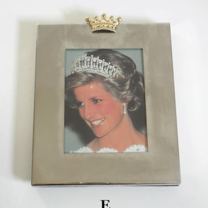 May include: A silver picture frame with a gold crown on top. The frame holds a photo of a woman wearing a tiara and a white dress. The letter "E" is printed below the frame.