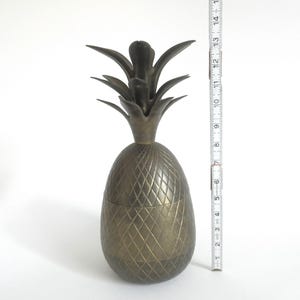May include: A gold-toned metal pineapple-shaped container with a textured surface. The container is approximately 15 inches tall.