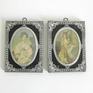 May include: Two ornate silver frames with black interiors, each containing a vintage portrait of a woman in a dress. The frames have intricate designs and are hung on a white wall.