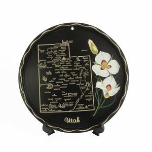 May include: A black metal round plate with a map of Utah and a white flower design. The plate has a black stand and the text "Utah" is printed on the bottom.