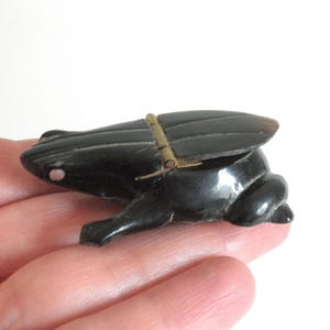 May include: A small, black frog figurine with a hinged, wing-shaped lid. The frog has a glossy finish and a small, white dot on its side. The wings are attached with a brass-colored hinge.