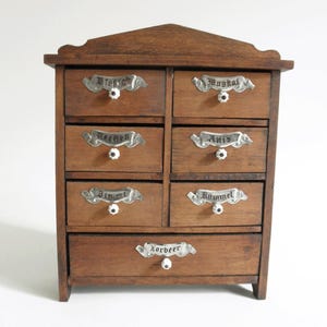 May include: A small, antique wooden spice cabinet with eight drawers. Each drawer has a decorative metal label and a white knob. The cabinet is made of dark wood and has a triangular top.