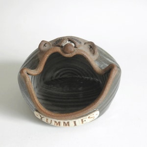 May include: A ceramic bowl shaped like a smiling face with the word "Yummies" written on the front. The bowl is glazed in shades of brown and gray.