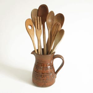 May include: A brown ceramic pitcher with a handle filled with wooden kitchen utensils, including spoons, spatulas, and a fork.
