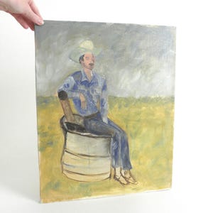 May include: An oil painting on canvas depicting a figure wearing a light-colored cowboy hat and blue clothing, seated on a cylindrical metal drum. The background features a yellow field under a grey, overcast sky, rendered in visible brushstrokes.