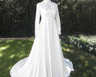 Könnte beinhalten: Ein weißes Vintage-Brautkleid mit langen Ärmeln und einem hohen, mit Spitze besetzten Ausschnitt. Das Kleid hat einen fließenden Rock und eine Schleppe, präsentiert auf einer Schaufensterpuppe in einer grasbewachsenen Umgebung. Klassischer Brautstil.