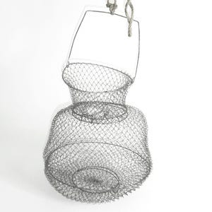 May include: A vintage metal wire fish basket with a handle and rope. The basket is cylindrical with a flared base and a smaller opening at the top. The wire is a dull silver color, and the basket is suspended against a white background.