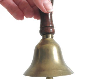 May include: A brass bell with a wooden handle. The bell is being held by a hand.